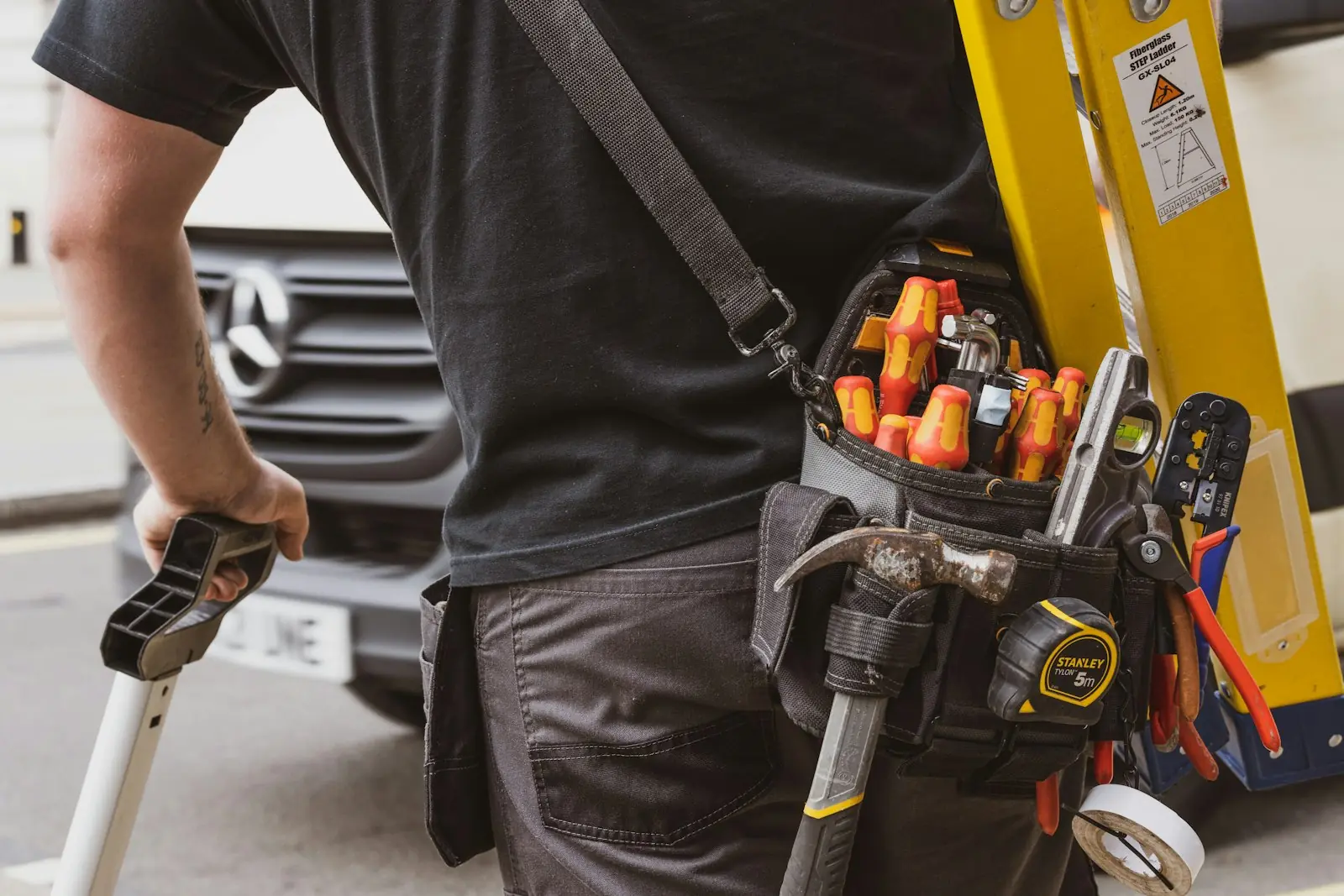 Technician with a tool belt of screwdrivers, hammer and tape measure, carrying a step ladder